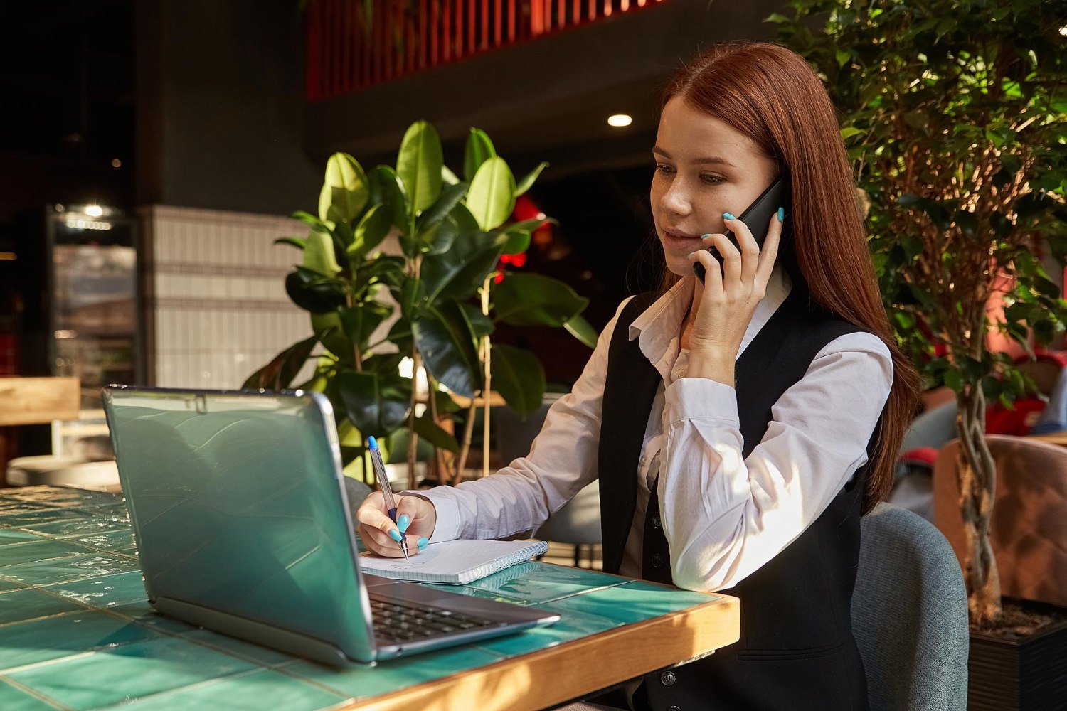 Girl student sitting at cafe table studying with laptop calling by smartphone